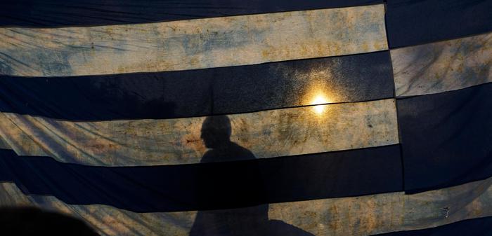 A man is silhouetted as he stands behind a representation of the the Greece national flag, as he takes part in a protest held by supporters of the NO vote in the upcoming referendum, at Syntagma square in Athens on Monday, June 29, 2015. Anxious Greek pensioners swarmed closed bank branches and long lines snaked outside ATMs as Greeks endured the first day of serious controls on their daily economic lives ahead of a July 5 referendum that could determine whether the country has to ditch the euro currency and return to the drachma. (ANSA/AP Photo/Daniel Ochoa de Olza)
